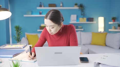 Asian-young-woman-working-from-home-both-taking-notes-and-working-from-laptop.